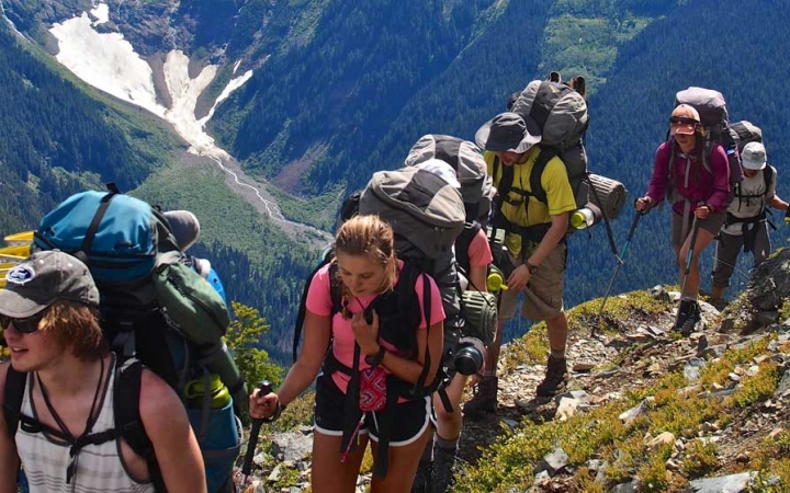 A group of students wearing backpacks hike along a trail high above a mountainous valley containing snow.